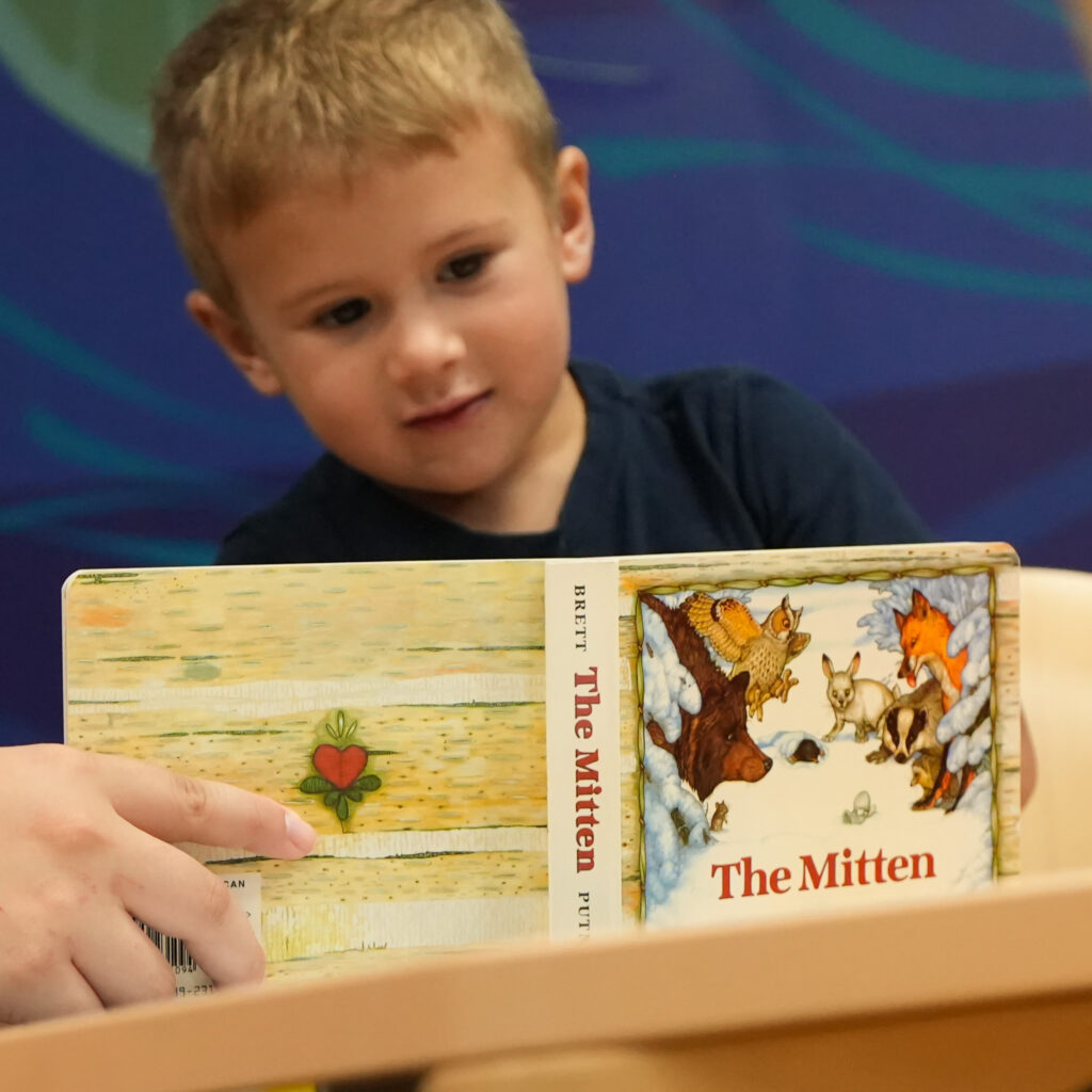 A young boy looks at a copy of "The Mitten." An adult holds the book for him to see. 